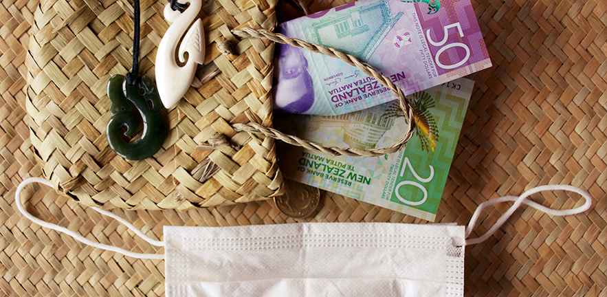 A mixture of New Zealand Bank notes with a face mask and a woven flax kete purse with Pounamu greenstone pendant. This image is for a Healthcare & Finance Concept.